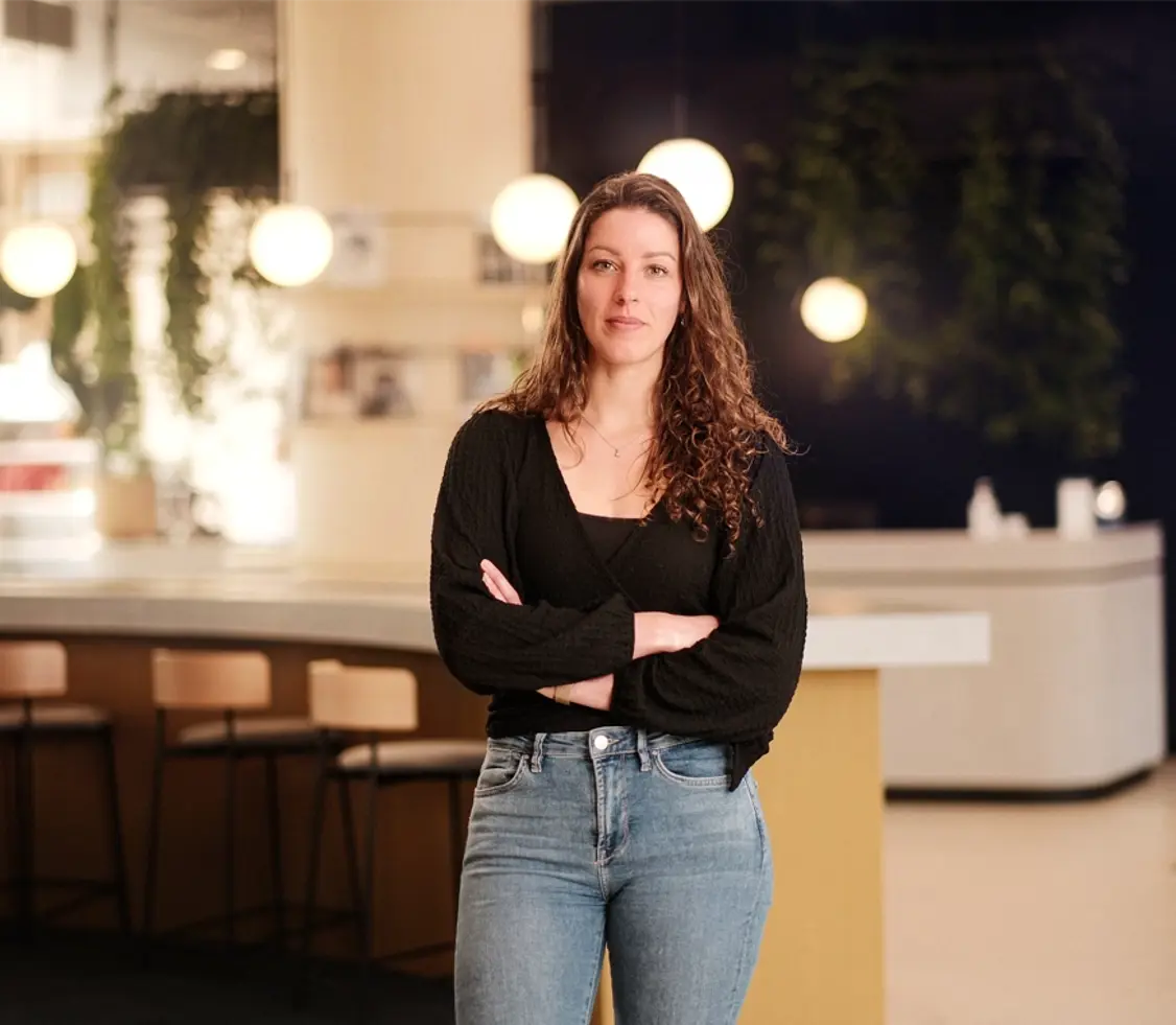 Jonge vrouw met bruin lang haar staat met gekruiste armen en kijkt lachend in de camera met op de achtergrond een Persoonality vestiging