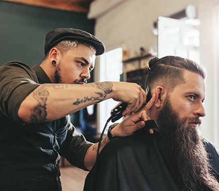 Een man is een andere man aan het scheren. De man links heeft tattoo's en de man rechts een lange baard